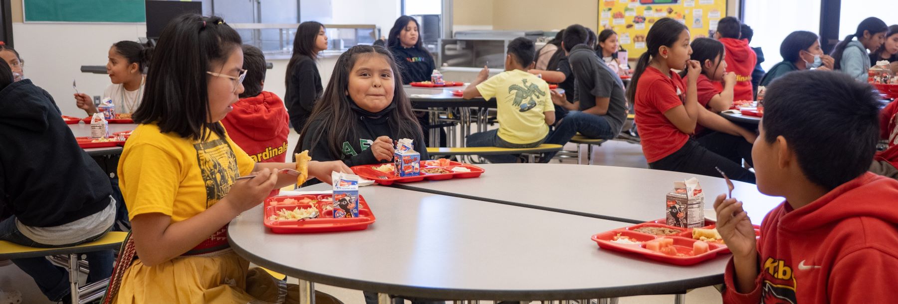 Students gathered in the school cafeteria, eating lunch and engaging in conversation at their tables.