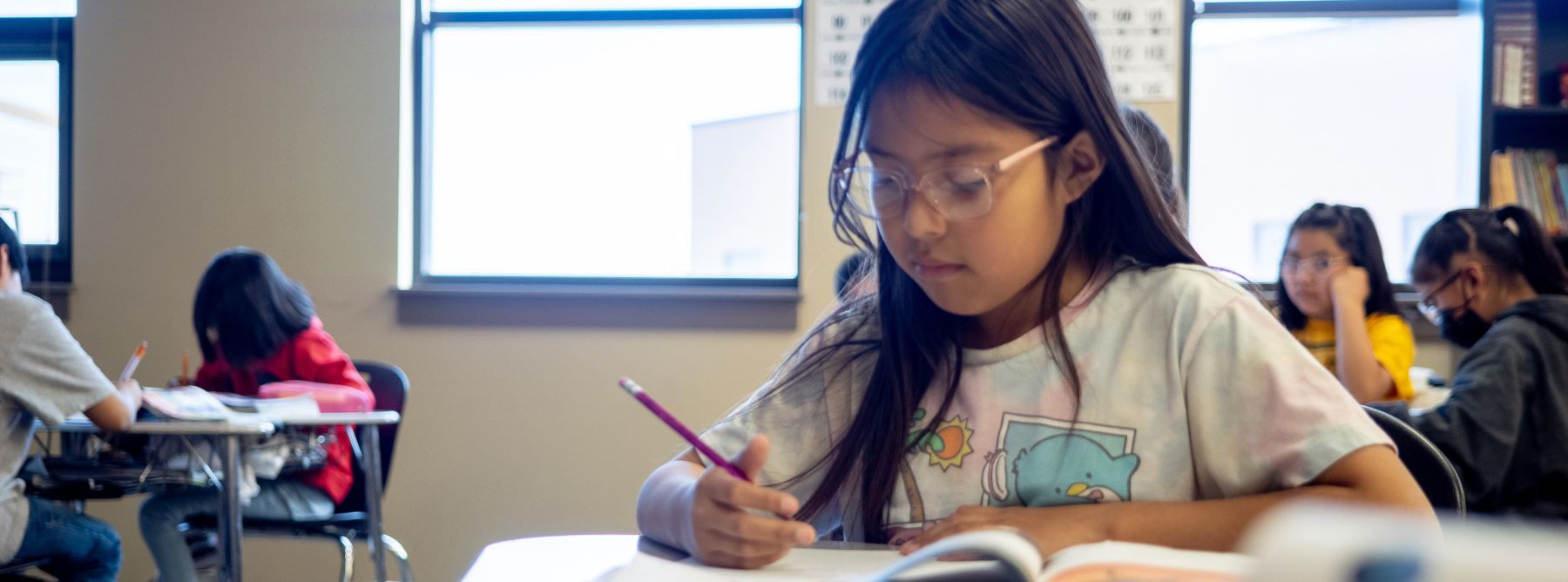 A young girl sits at a desk in a classroom, focused on her assignment.