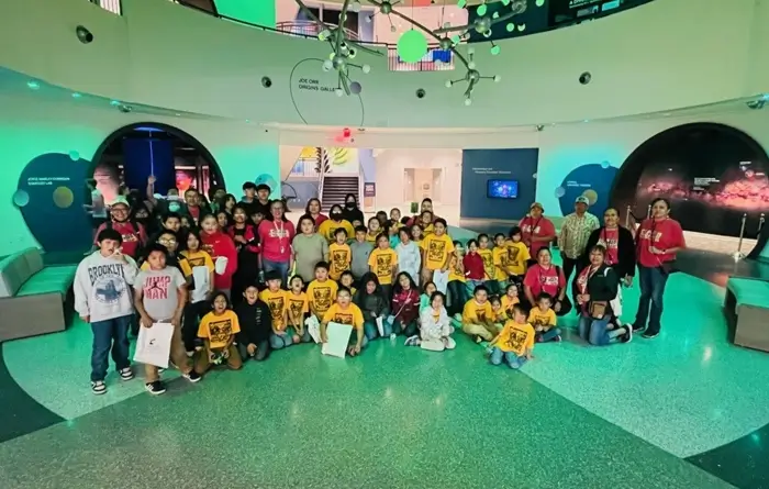 A large group of children and adults pose together for a photo inside a science museum or planetarium, standing under a ceiling display of green and white hanging spheres. Some children wear matching yellow T-shirts with a logo, while others wear casual clothing.  